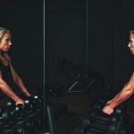 woman wearing black top top holding black dumbbells standing in front of mirror