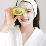 Smiling woman with face mask holding avocado for skincare in spa setting.