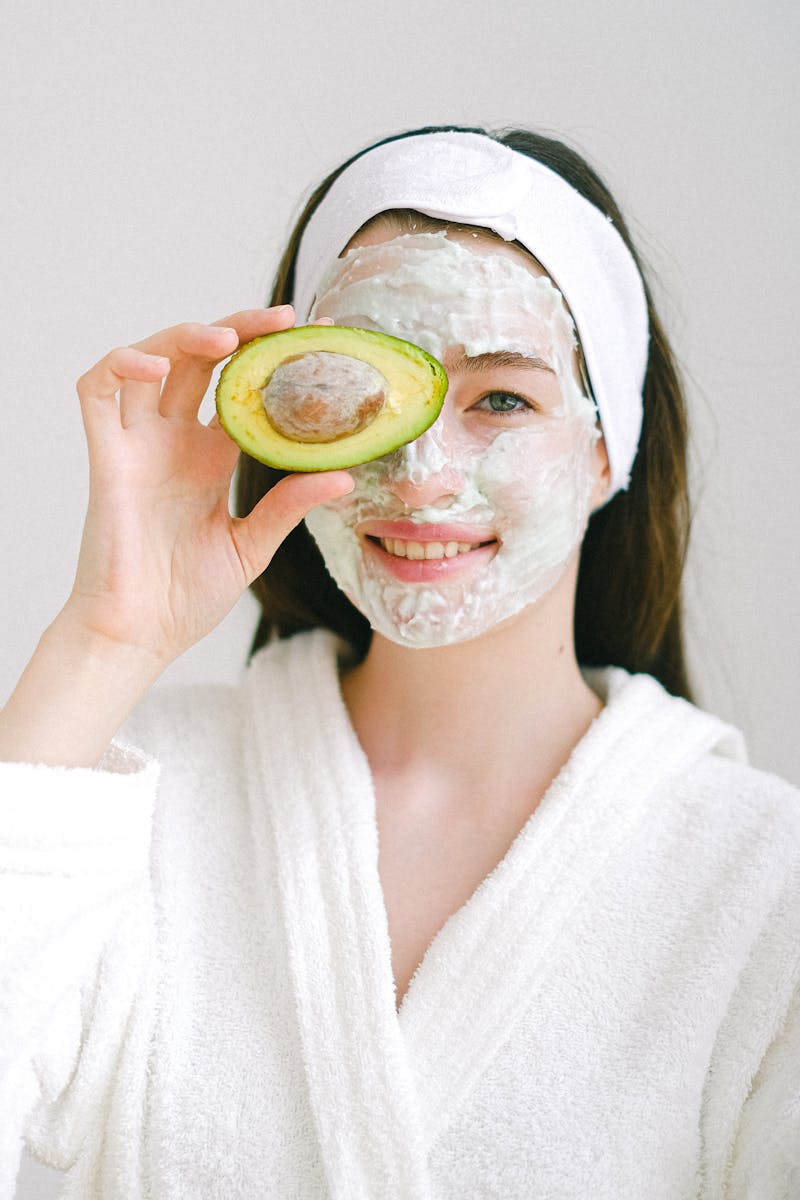 Smiling woman with face mask holding avocado for skincare in spa setting.